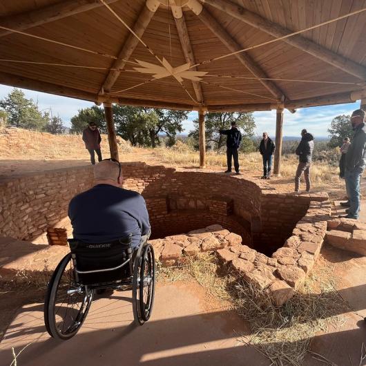 This photo shows an ancient kiva, or underground dwelling, which is shaded by a wooden canopy. A man in a wheelchair is looking into the kiva and six other people can be seen standing around the site. Pinon-juniper trees can be seen in the background.