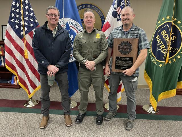 Three men pose for a photo in front of an American flag and a border patrol flag.